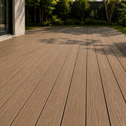Brown composite decking planks installed on an outdoor patio, with a modern house entrance on the left and green trees and shrubs in the background.