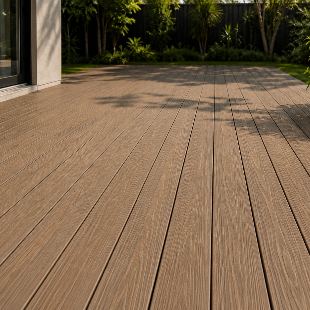 Brown composite decking planks installed on an outdoor patio, with a modern house entrance on the left and green trees and shrubs in the background.