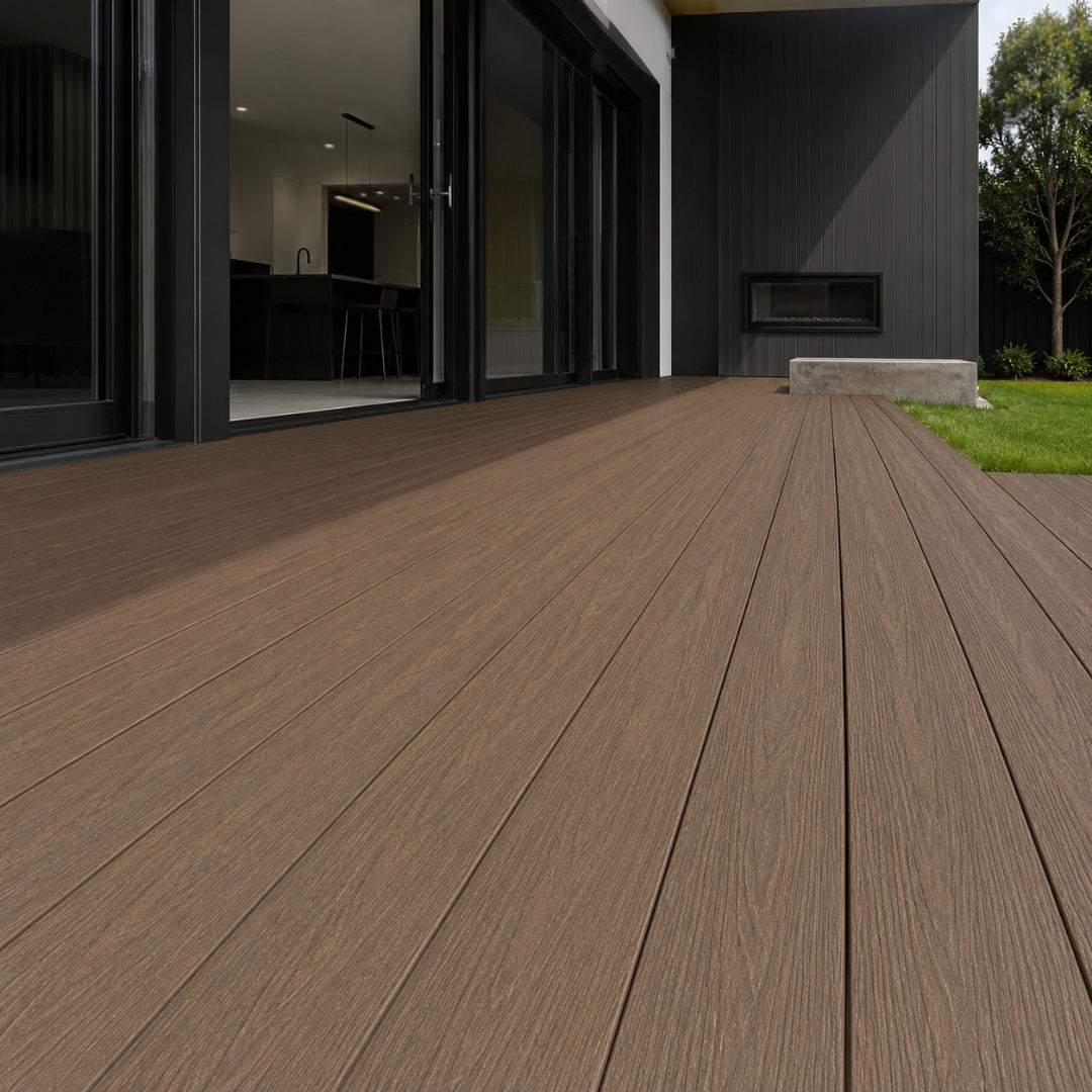 Brown composite decking planks installed on an outdoor patio in front of a modern house with black framed sliding glass doors and a small grassy area