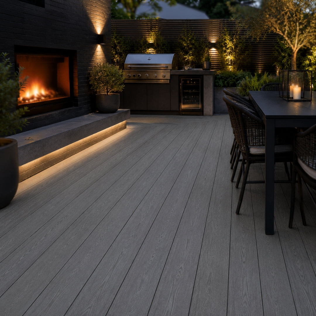 Black composite decking planks installed on a modern outdoor patio at dusk, shown with a built-in fireplace, grill, dining table, and warm ambient lighting.